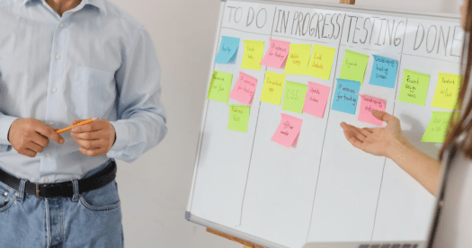 A man in a light blue shirt holds an orange pencil while standing next to a whiteboard covered with colorful sticky notes organized into 'To Do', 'In Progress', 'Testing', and 'Done' categories.