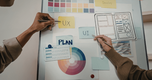 A close-up of two hands working on a design board, featuring sticky notes labeled 'UX' and 'UI,' a layout plan, a color wheel, and various design elements.