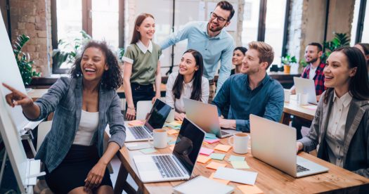 A diverse group of professionals engaged in a collaborative meeting, with one person presenting on a whiteboard while others smile and interact. Laptops and sticky notes are spread across the table in a bright, modern office setting.